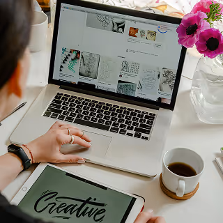 Person working on brand visuals using a laptop and a tablet labeled “Creative,” with sketches and flower vase on the desk, depicting a creative workspace.