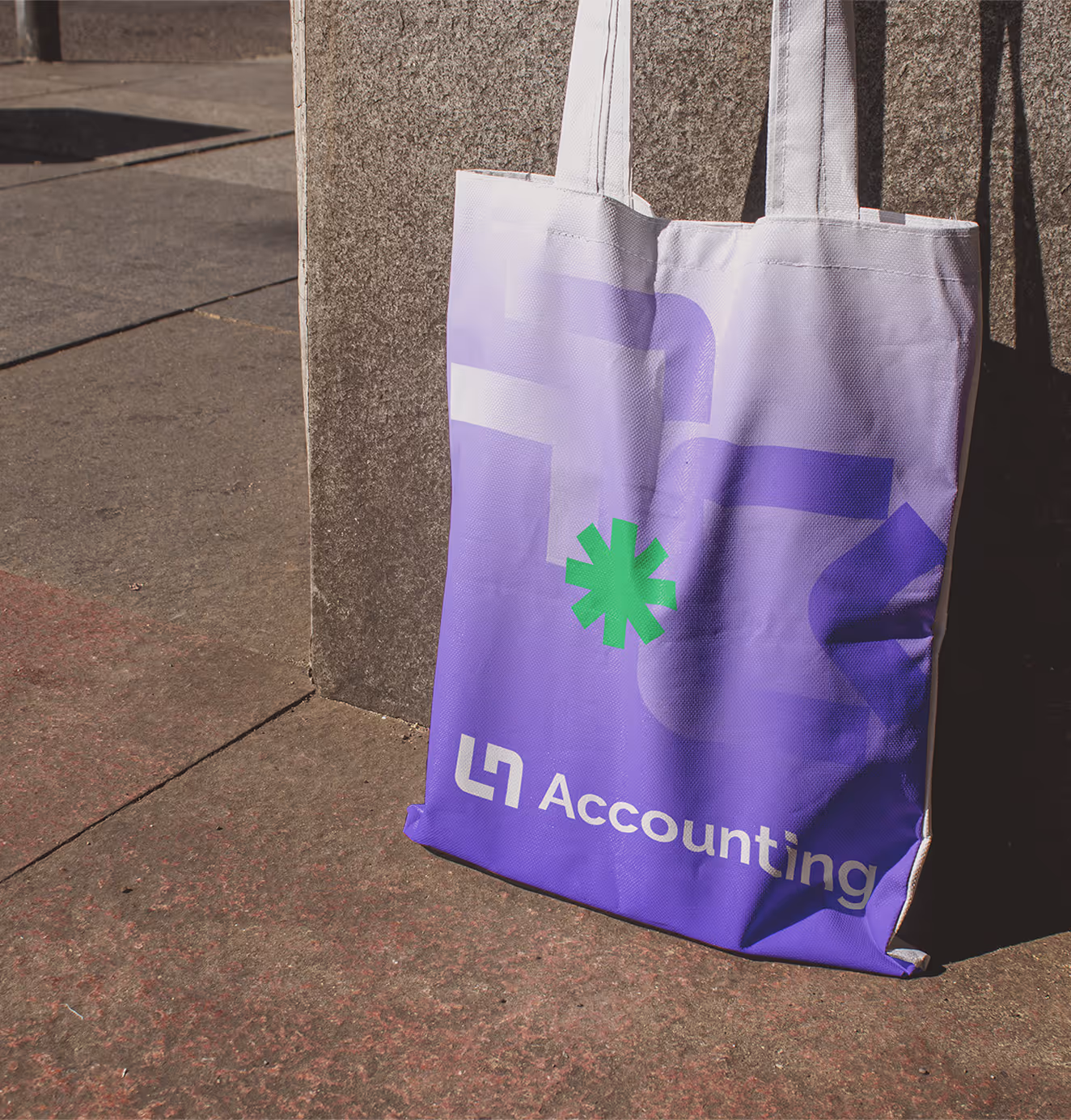 LN Accounting branded tote bag in a gradient purple design with green geometric star icon and bold white logo, leaning against a concrete wall outdoors.