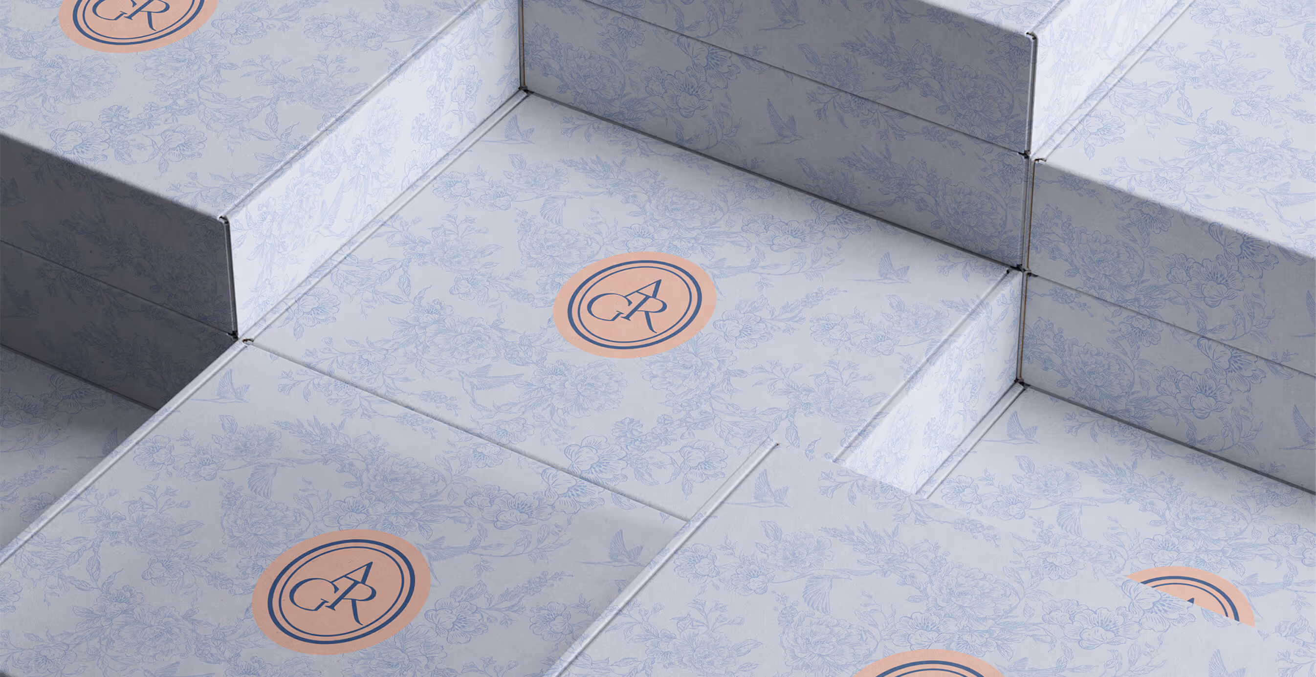 Elegant light blue boxes with floral patterns and a peach monogram logo stacked neatly, showcasing luxury packaging design.