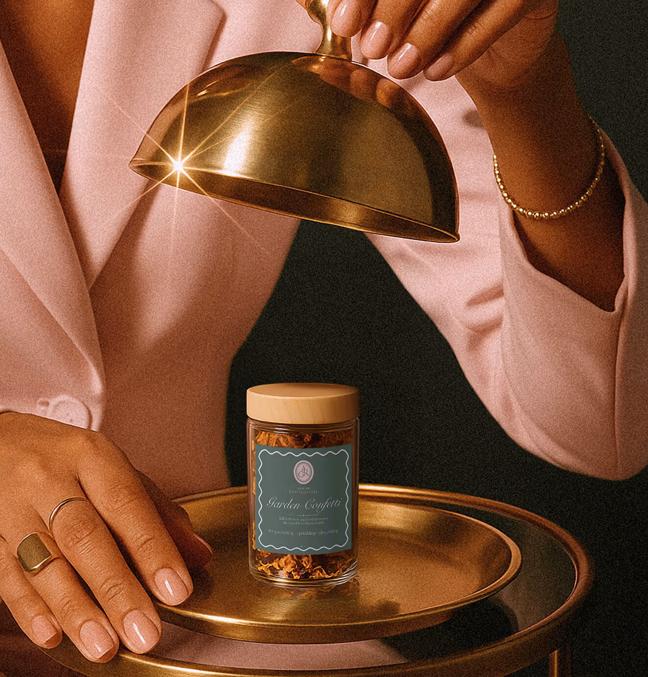 Close-up of a woman in a pink blazer lifting a gold cloche to reveal a premium jar labeled “Garden Confetti” on a golden tray.