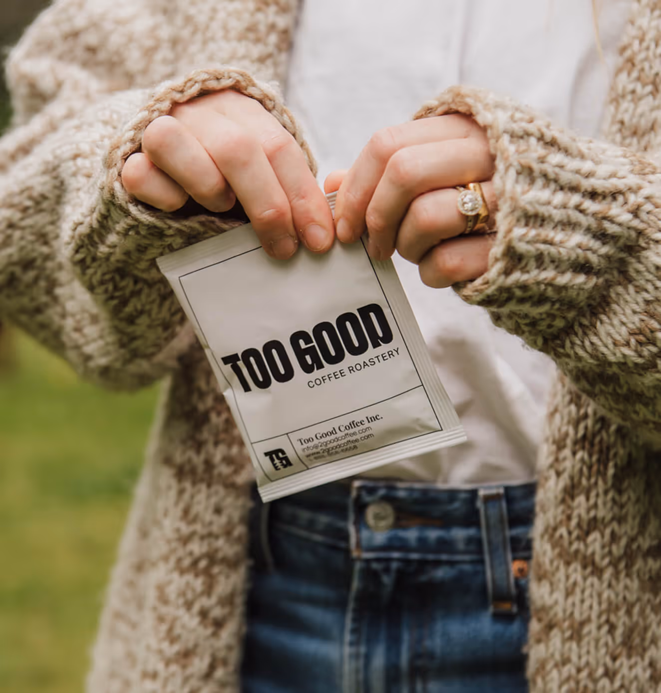 Close-up of a person in a cozy knit sweater holding a white Too Good Coffee Roastery sachet outdoors, with greenery in the background.