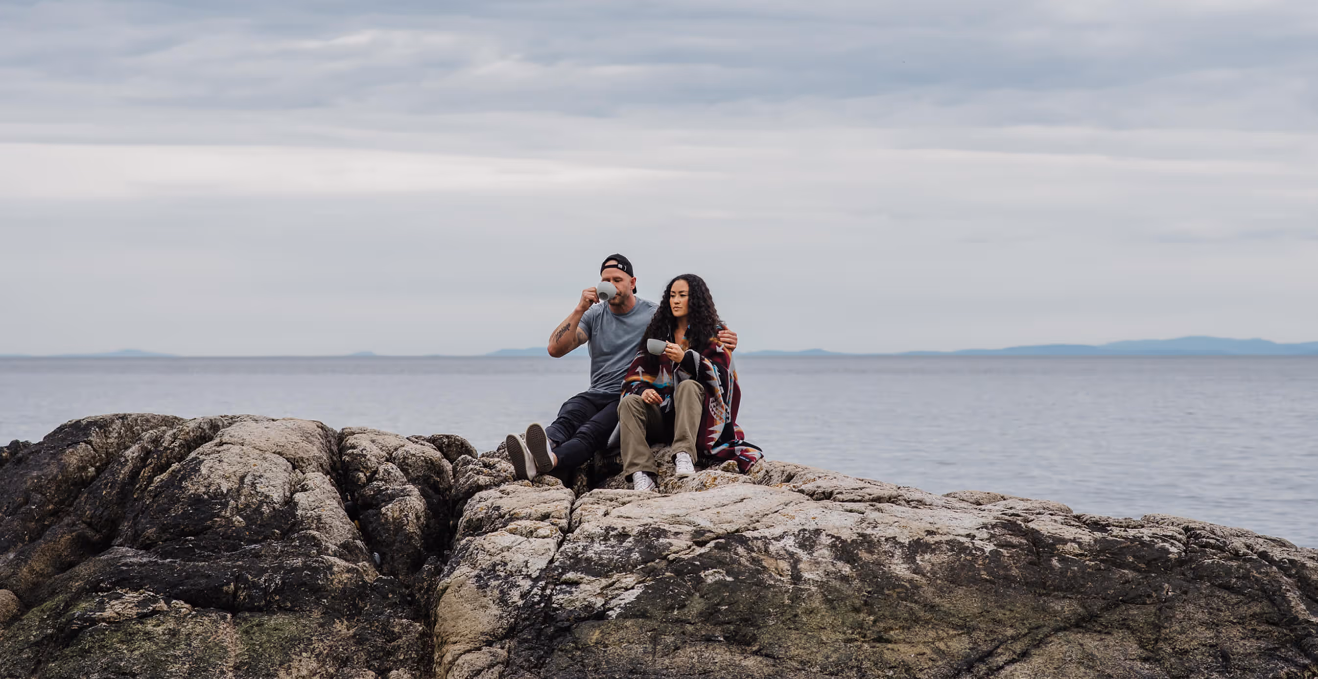 A couple sits on large seaside rocks, drinking coffee together. The man wears a cap and grey shirt, the woman is wrapped in a patterned blanket, both holding mugs with the ocean behind them.