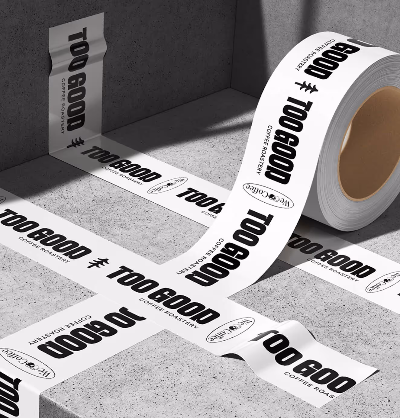 A roll of Too Good Coffee Roastery branded tape in black lettering on white, unrolled across a concrete surface in a cross pattern.