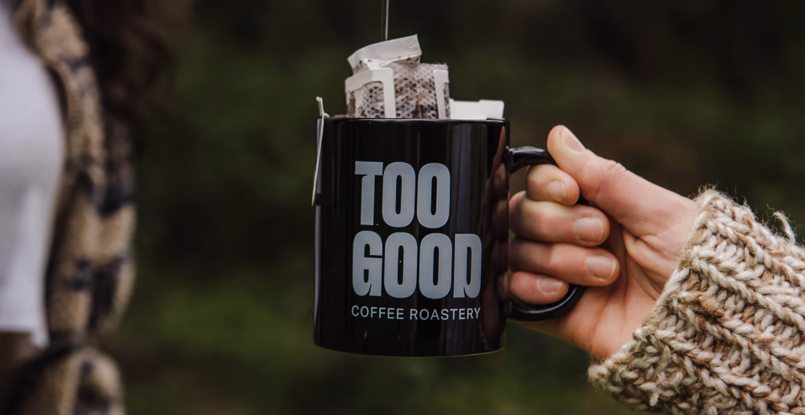 Close-up of a hand holding a black Too Good Coffee Roastery mug filled with drip coffee filters, with a blurred outdoor background.
