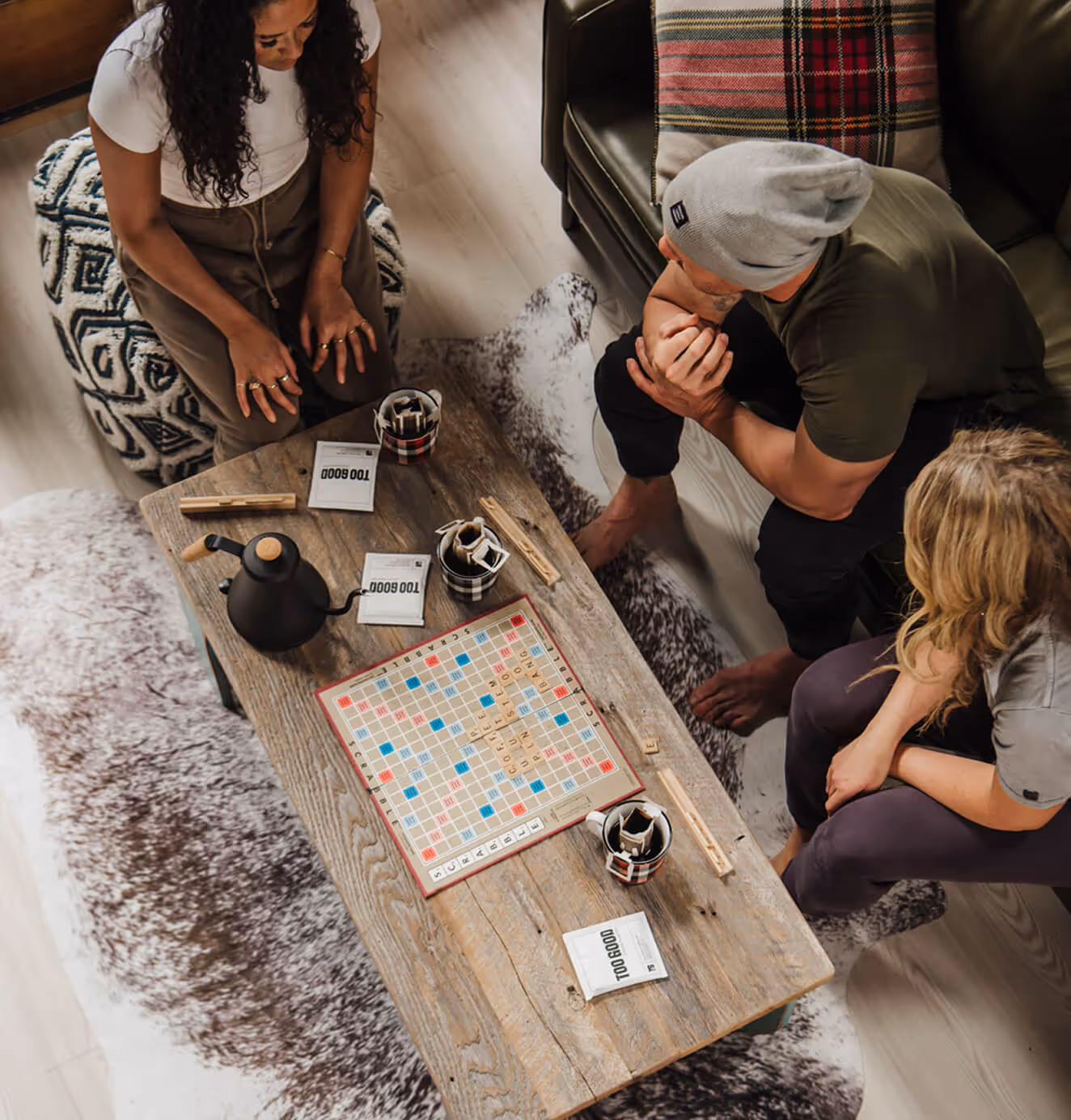 Overhead view of three friends sitting around a rustic wooden coffee table playing Scrabble, with Too Good Coffee Roastery packets and mugs on the table.