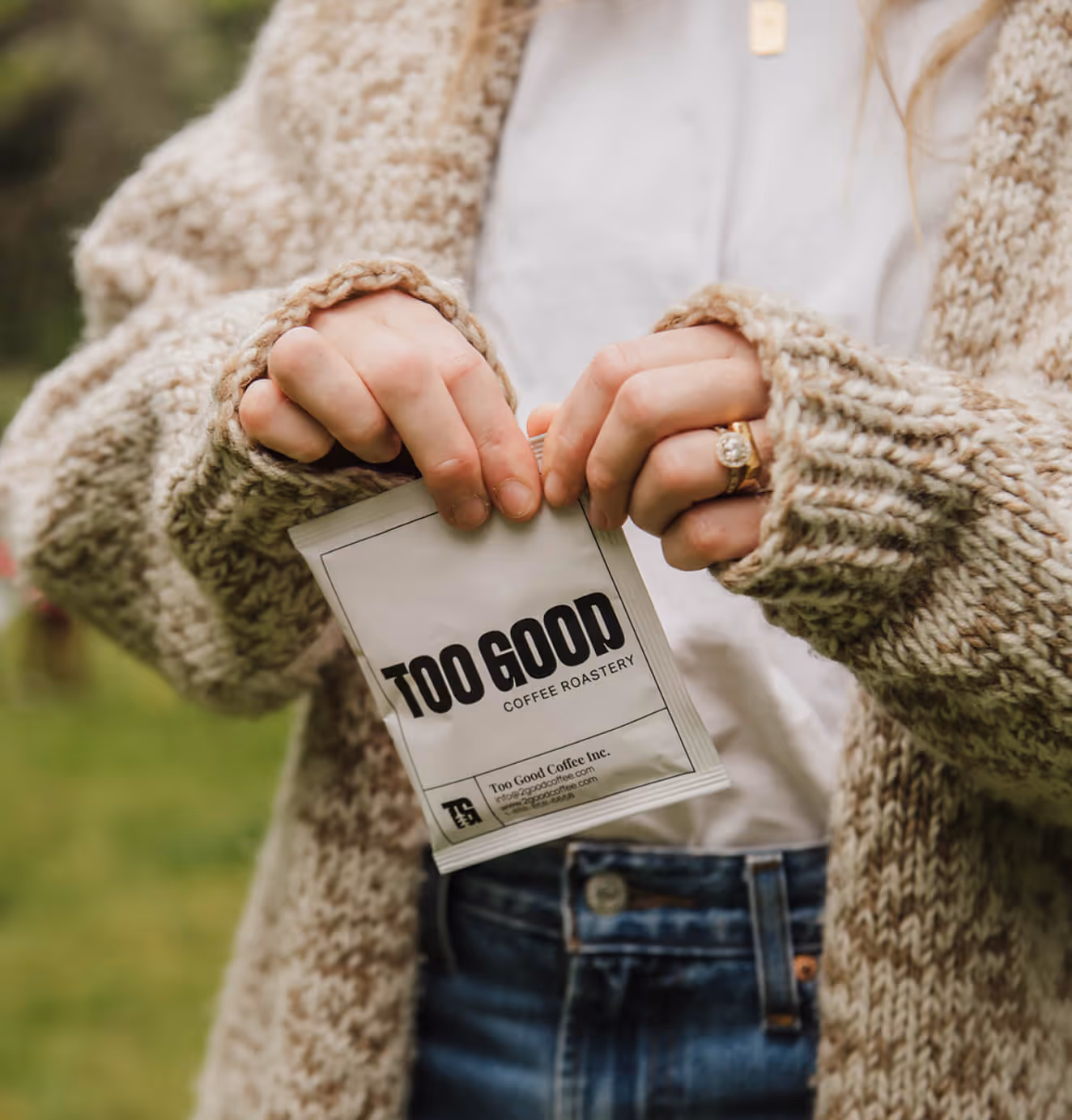Close-up of a person in a cozy knit sweater holding a white Too Good Coffee Roastery sachet outdoors, with greenery in the background.