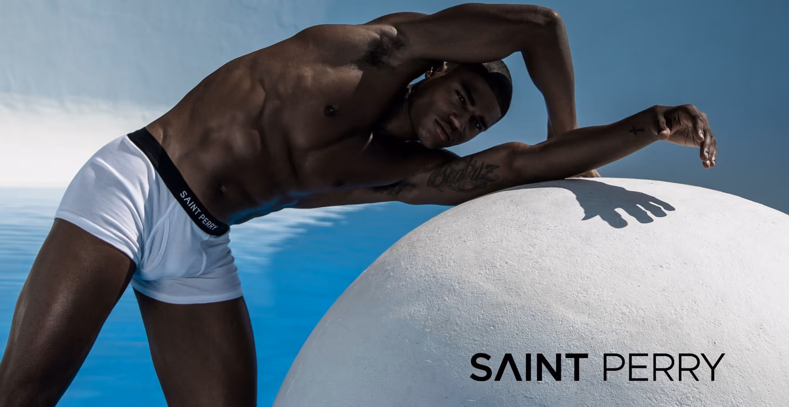 A male model poses shirtless in white Saint Perry underwear, stretching against a large white sphere with a blue water-inspired backdrop.