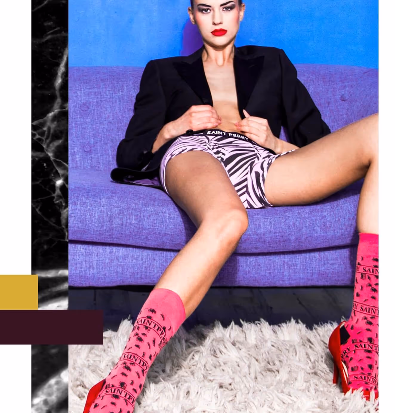 A female model reclines on a purple couch wearing a black blazer, zebra-print Saint Perry shorts, and pink branded socks with red high heels.