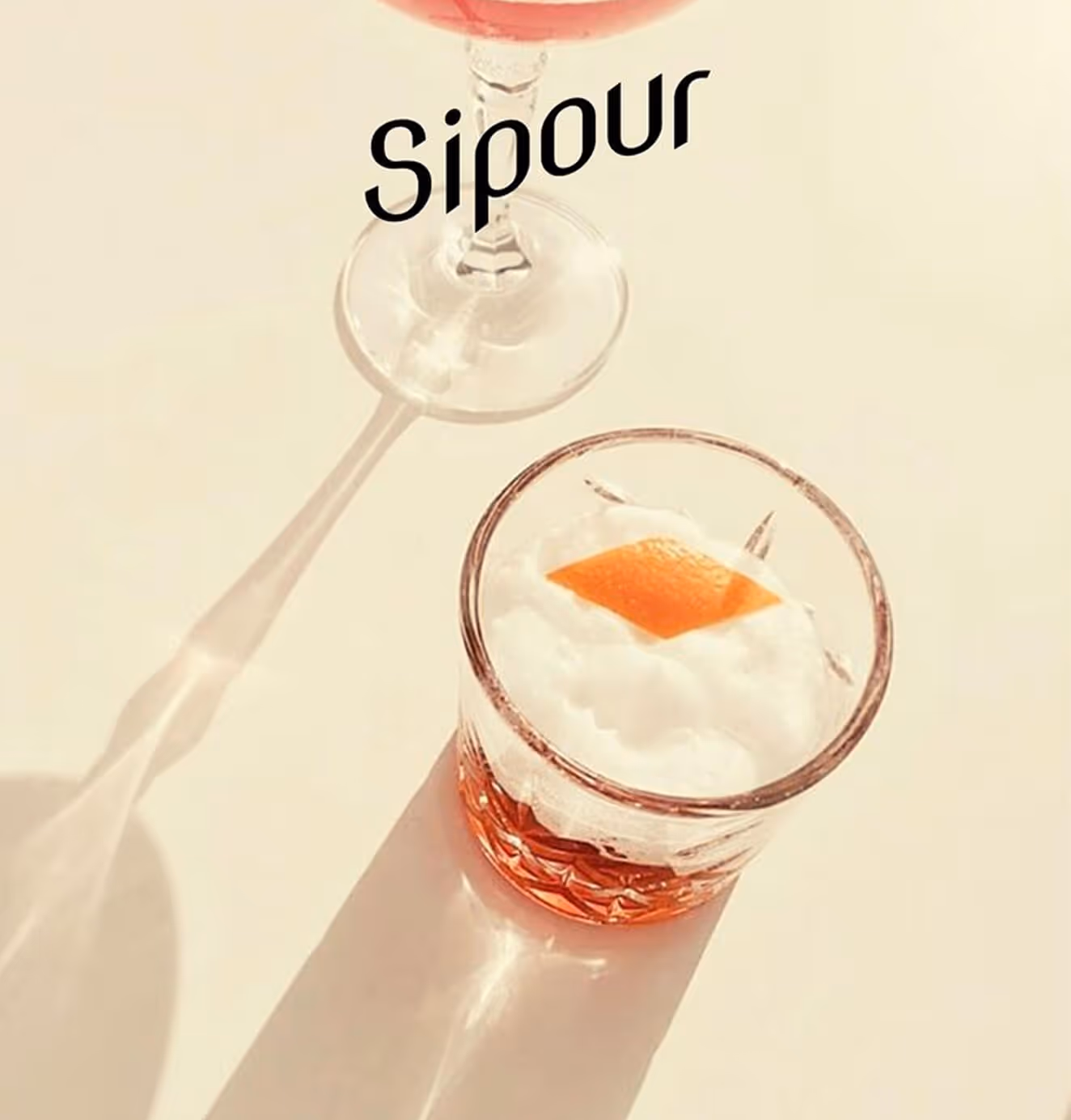 Two Sipour cocktails in elegant glassware on a sunlit surface, one with frothy foam and orange garnish, the other in a stemmed glass casting a long shadow.