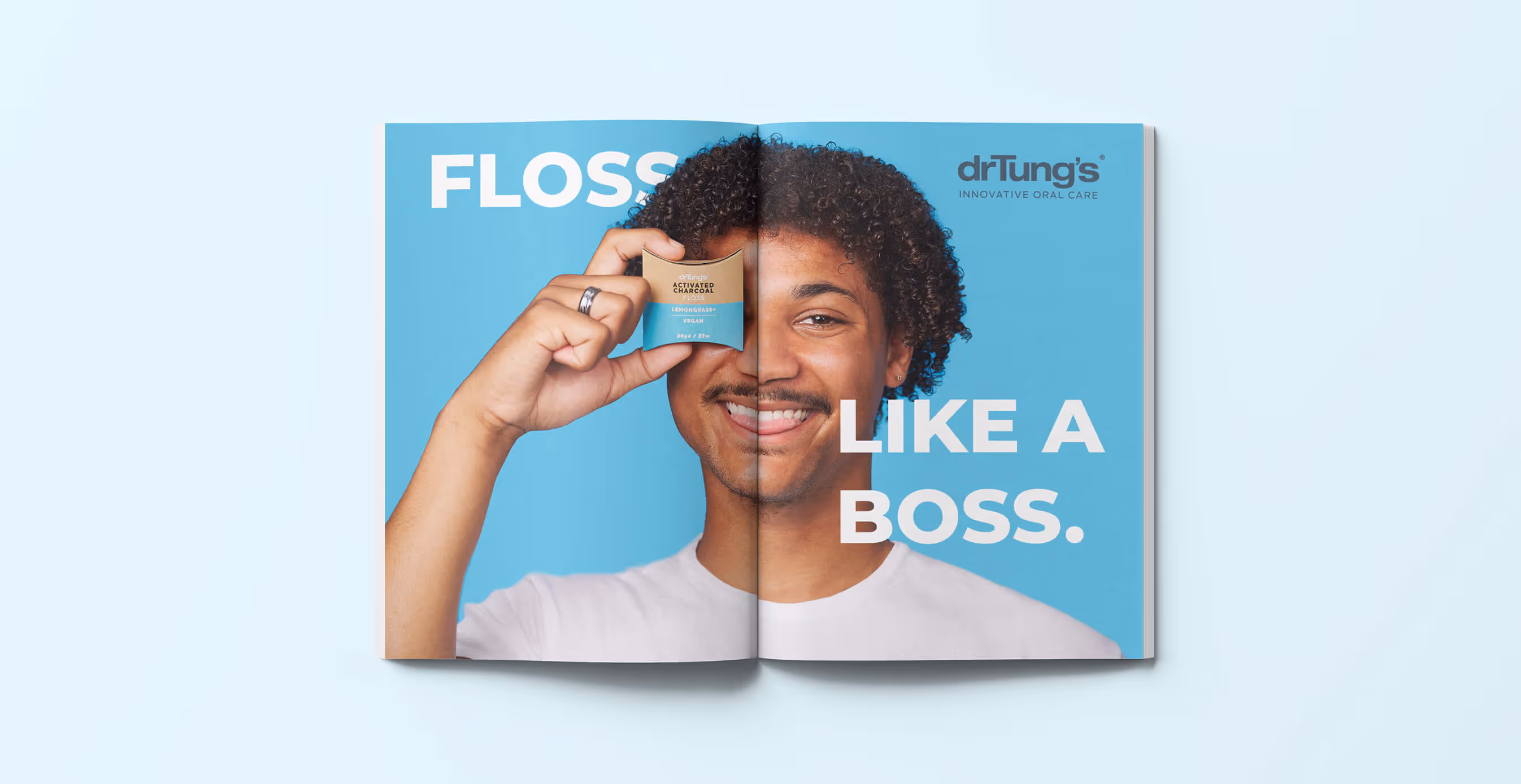 Magazine spread ad for Dr. Tung’s featuring a smiling man in a white shirt holding a box of Activated Charcoal Floss over one eye. The bold text reads ‘Floss Like a Boss’ against a bright blue background with Dr. Tung’s Innovative Oral Care logo.