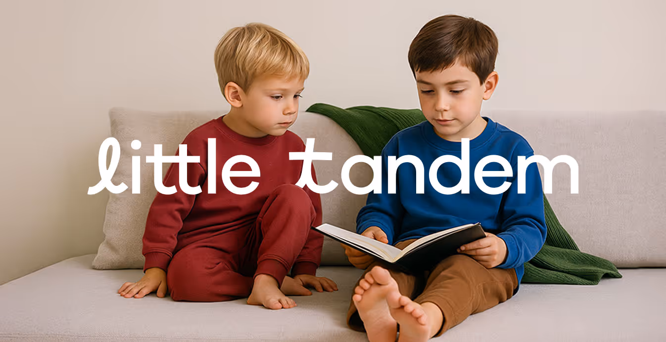 Two young boys sit together on a neutral sofa, one in red pajamas and one in blue, reading a book with the Little Tandem logo overlay.