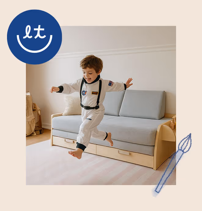 A young boy dressed in a white astronaut costume joyfully jumps on a beige modular sofa with built-in drawers, smiling in play.