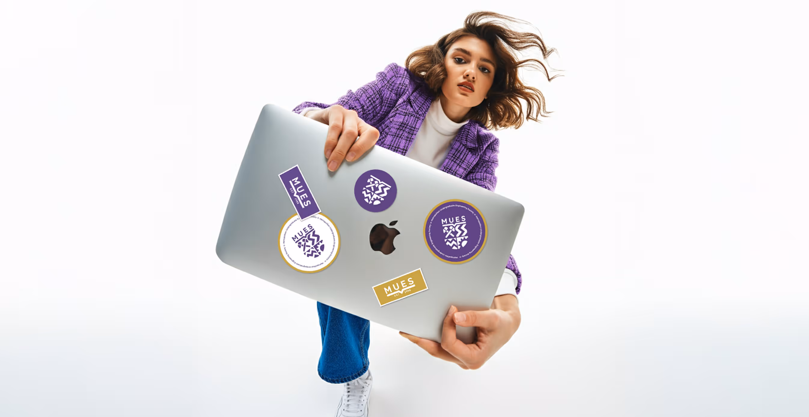 A student in a purple jacket holds a laptop decorated with multiple MUES logo stickers, including circular and rectangular designs in purple, gold, and white.