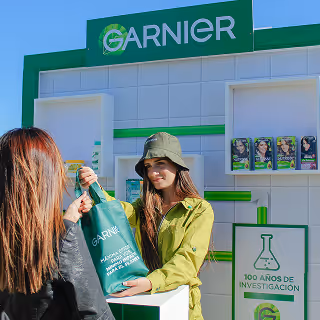 Brand ambassador at a Garnier promotional stand handing a tote bag to a passerby, highlighting a live marketing activation with product displays.