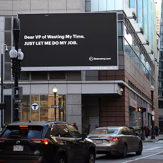 Outdoor billboard on a city building reads, 'Dear VP of Wasting My Time, JUST LET ME DO MY JOB.' by Basecamp, showcasing bold offline advertising.