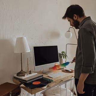 Man cleaning a home office desk with a feather duster, next to a computer, lamp, and record player, representing workspace organization.