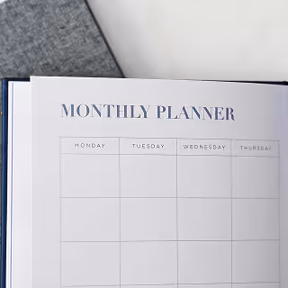Open monthly planner showing a blank calendar layout for Monday through Thursday, placed on a light surface with a fabric notebook beside it.