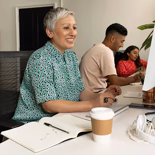 Smiling woman with short gray hair in a patterned green shirt working at a desk with a coffee cup, notebook, and computer, alongside two colleagues.