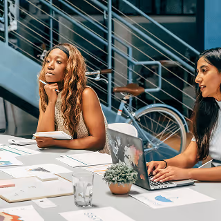 Two women sitting at a table in a modern office space, attentively listening and working on marketing strategy documents with charts and a laptop.