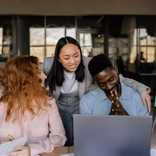 Three diverse coworkers happily collaborating at a desk, with one woman standing and engaging the others as they look at a laptop screen.