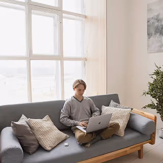 Young man sitting cross-legged on a modern gray sofa, working on a laptop near a large window with natural light.