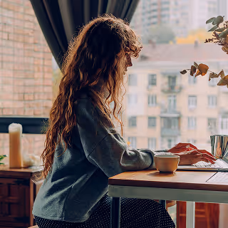 A woman with long curly hair sits at a desk by a window, typing on a keyboard with a coffee cup nearby. City buildings are visible through the window, creating a cozy urban work-from-home setting