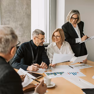 Four older professionals in business attire sit around a table discussing documents with charts and graphs, while a woman standing nearby takes notes on a clipboard. They appear to be in a bright, modern office setting.