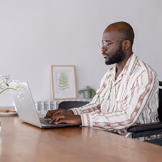 A man with glasses and a patterned shirt types on a laptop at a wooden desk, with a framed plant print and a vase in the background, suggesting a calm, minimalist workspace.