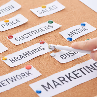 Close-up of a corkboard with white paper labels pinned in place, showing words like "BRANDING," "MARKETING," "CUSTOMERS," and "SALES," with a hand pointing at one using a pencil.