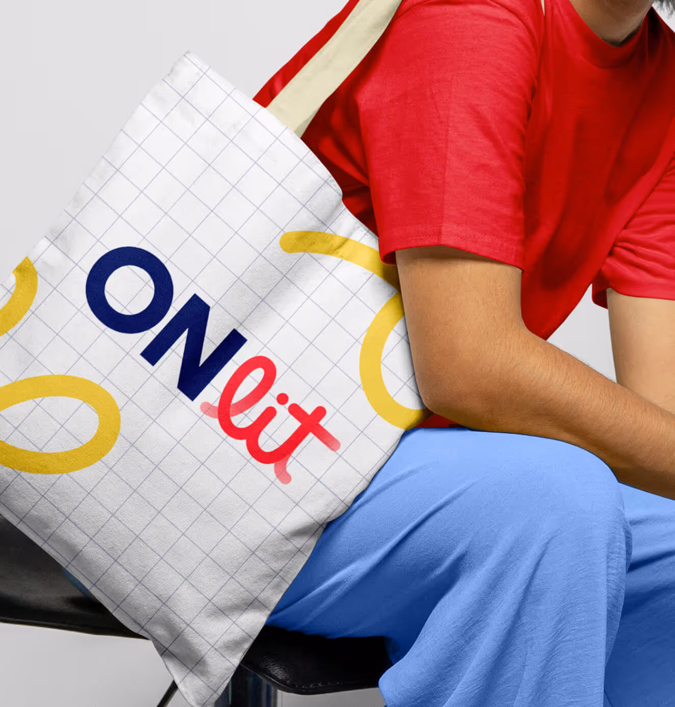 Person sitting with a white tote bag featuring ONlit logo in navy and red, a yellow looping graphic, and a light grid background.