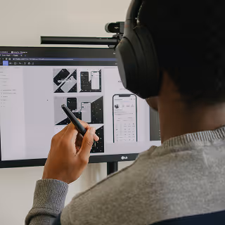A person wearing headphones and using a stylus to review black-and-white mobile app wireframes on a computer screen.