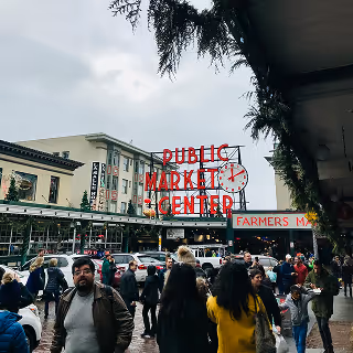 Crowd of people walking near Seattle’s iconic Public Market Center sign on a cloudy day, with the Farmers Market entrance visible.