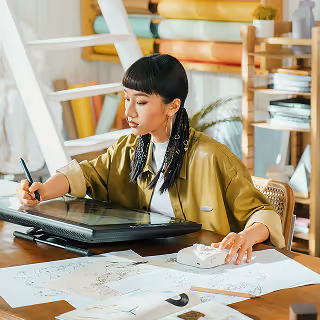 Young woman using a digital drawing tablet in a bright, creative workspace, surrounded by sketches and art supplies.