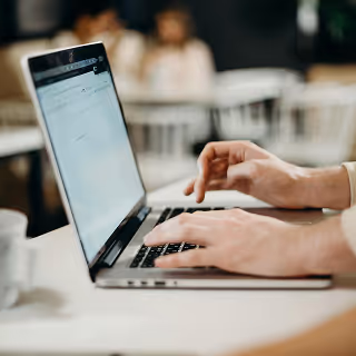 Close-up of a person typing on a laptop at a white table in a cozy café setting, with a blurred background of people sitting and chatting.