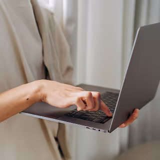 Partial view of a person holding and typing on a slim laptop while standing in a softly lit room with light curtains in the background.