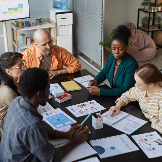 A diverse team of five professionals collaborating around a table covered in printed marketing charts and notebooks in a bright, modern office.
