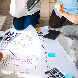 A group of people kneeling around a large white poster paper with hand-drawn diagrams, notes, and sticky notes during a creative planning session.