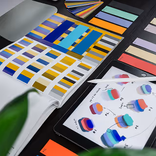 Desk setup with printed color palettes, a tablet showing brand color experiments, and various creative tools, representing the brand development process.