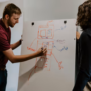 Two people collaborating at a whiteboard filled with handwritten diagrams and notes, discussing content strategy and event planning.
