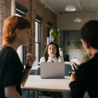 Three coworkers in a casual meeting; one woman looks thoughtful while sitting at a laptop, with two colleagues in conversation in the foreground.