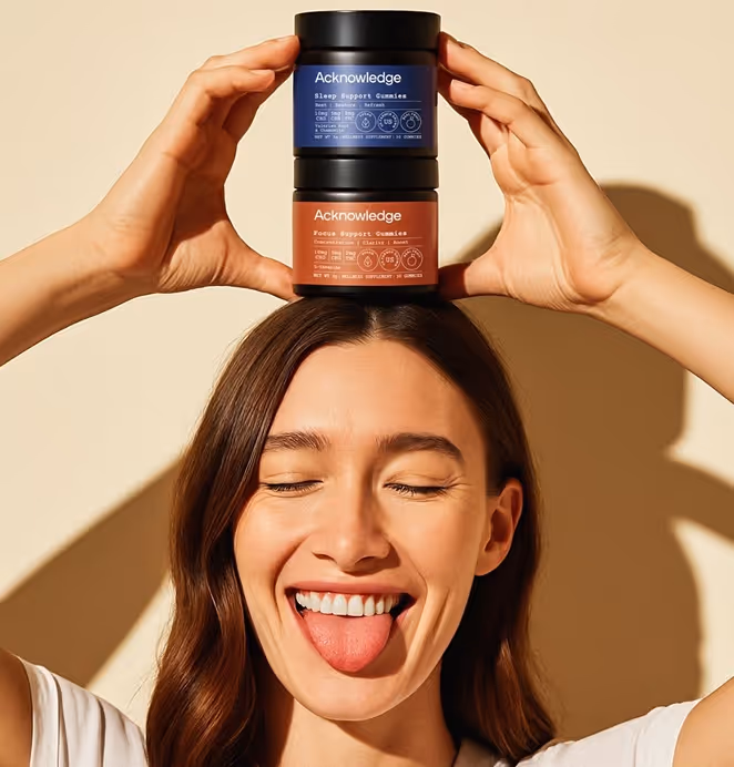 A smiling woman with long brown hair playfully sticks out her tongue and closes her eyes while holding two stacked jars of Acknowledge brand gummies on top of her head. The top jar has a blue label labeled “Sleep Support Gummies,” and the bottom jar has a rust-red label labeled “Focus Support Gummies.” The background is a warm beige, and the lighting creates soft shadows, giving the image a cheerful and vibrant feel.