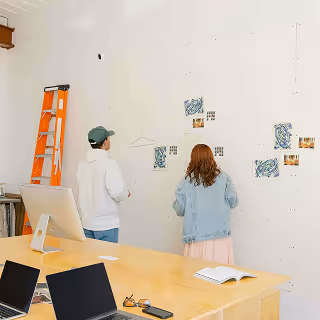 Two creatives stand in front of a white wall reviewing mood board pieces and sketches, collaborating in a minimalist studio environment with an orange ladder nearby.