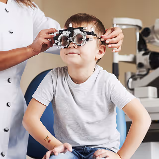 Smiling young boy undergoing an eye test with a trial frame, assisted by an optometrist in a white coat.