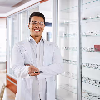 Friendly optician standing in front of a glass display of eyeglass frames, holding a pair of glasses with a welcoming smile.