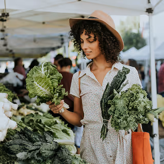 A woman in a hat shopping for leafy greens at an outdoor farmer's market, highlighting local, eco-conscious produce vendors.