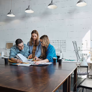 Group of three professionals sitting at a large wooden desk in a studio-like space, collaborating on printed materials and design layouts.