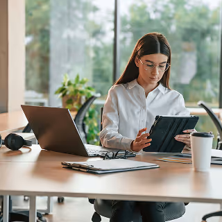 Professional woman wearing glasses and white blouse, sitting at a modern office desk using a tablet, with a laptop, coffee cup, and plants in the background.
