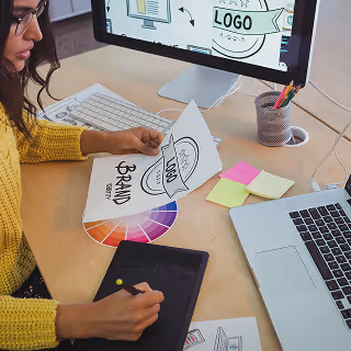 A designer at a desk reviewing a brand identity sketch, surrounded by color swatches, a computer screen showing logo concepts, and digital tools.