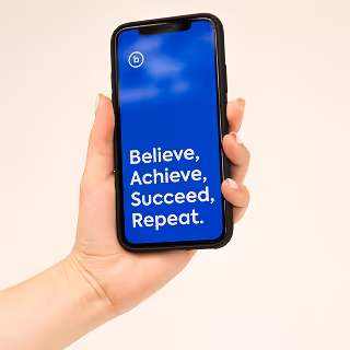 A hand holding a smartphone displaying a bold blue screen with motivational branding text: “Believe, Achieve, Succeed, Repeat.”