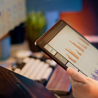 Hand holding a tablet displaying bar graphs and data charts, with a blurred office setup in the background.